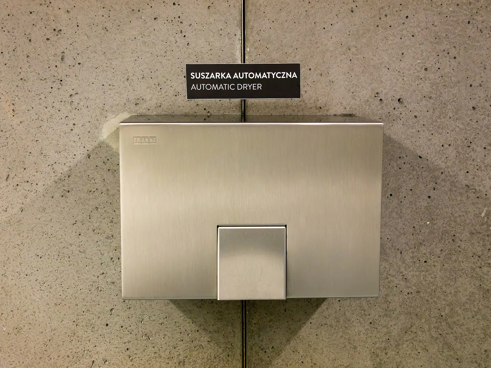 Händetrockner aus gebürstetem Edelstahl an einer Betonwand in einem öffentlichen Waschraum